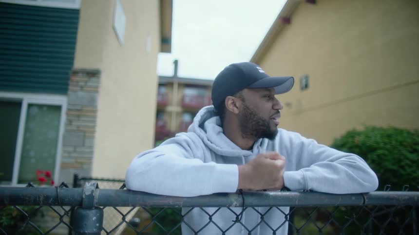 A man stands outside an apartment building and taps his hand on a fence as he looks around