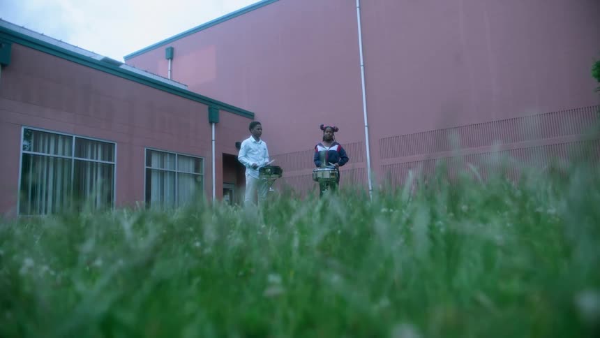 A boy and girl stand outside a school and drum