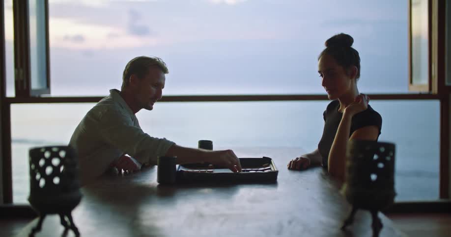 Man and woman sit at table playing a board game near a window showing the ocean