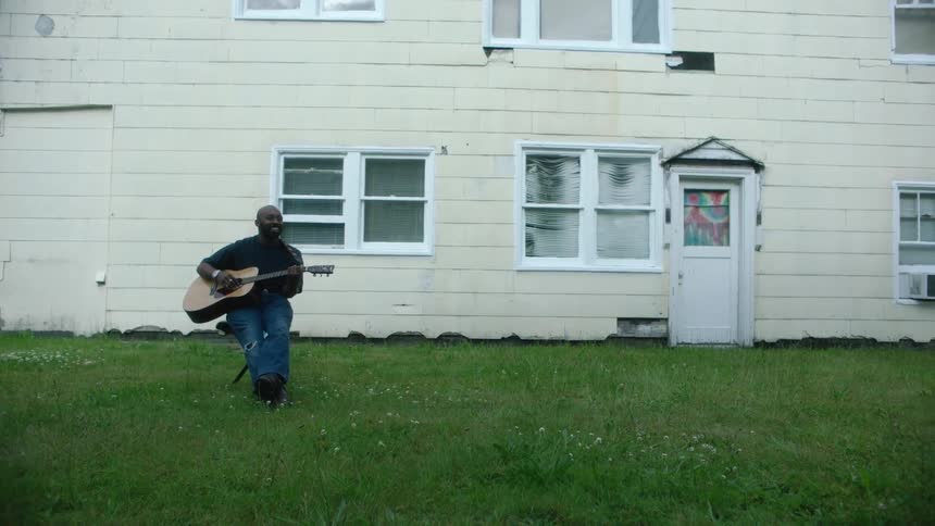 Man sitting on a chair in the grass in front of a house playing acoustic guitar