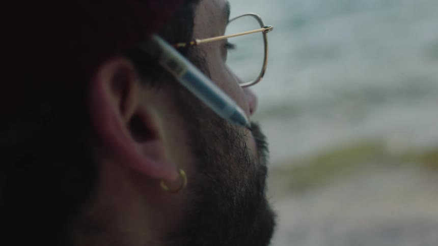 A young bearded man wearing glasses looks out upon the ocean as waves wash ashore