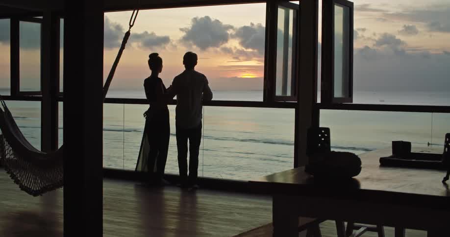 Silhouette of a young couple holding hands walking towards the window of a waterfront apartment at sunset