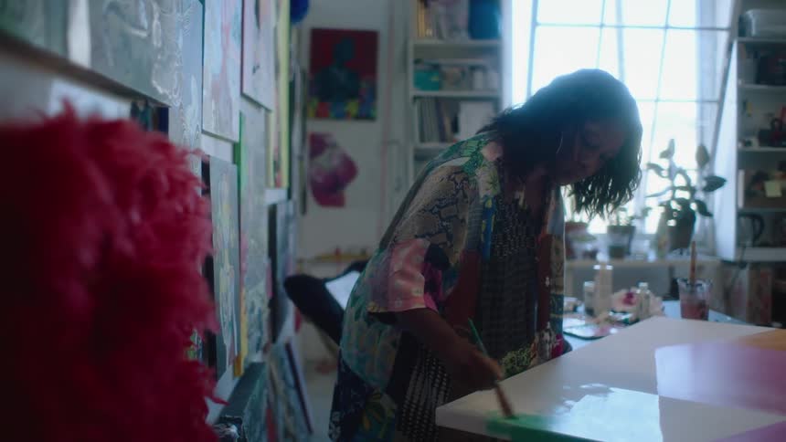 Female painter with wavy hair hoop earrings and shirt paints picture in room with pictures on walls