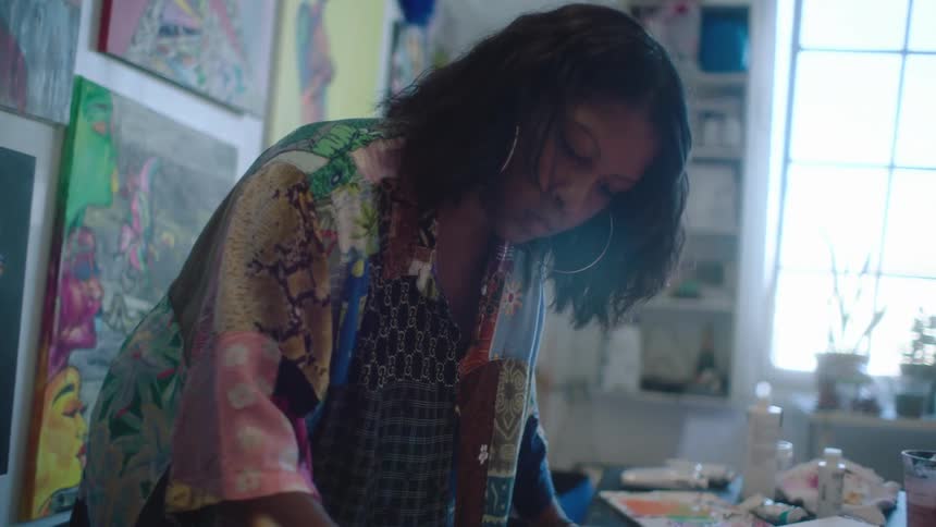 Female painter with wavy hair hoop earrings and shirt paints picture in room with pictures on wall