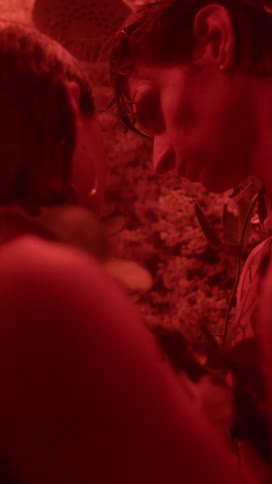 A man and a woman are standing in a room filled with plants and he is looking into her eyes