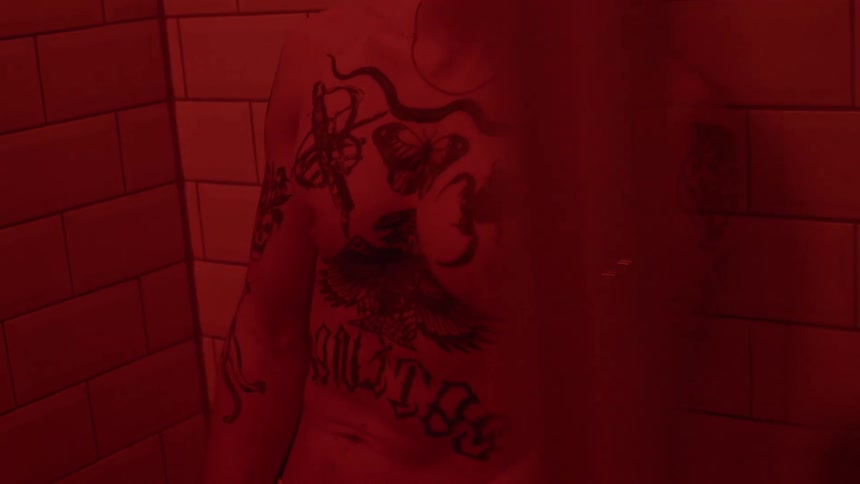 A man covered in tattoos on his chest and arms is leaning up against a subway tile wall in the red lighting