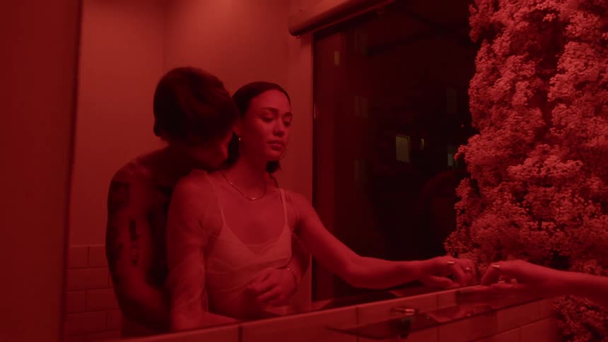Man back hugs woman as they stand in bathroom under red colored lighting in front of mirror