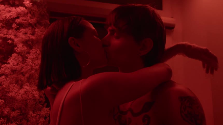 A woman puts her arms around a mans neck and kisses him and he looks at her with a smile in the glow of a red light