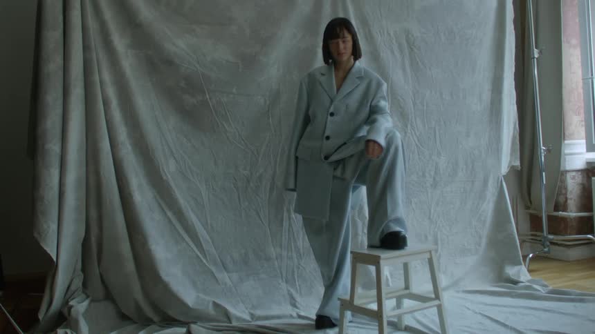 A woman is standing in a room with her foot on a stool modeling oversized clothes that are clearly way too big for her and there is a sheet on the wall behind her