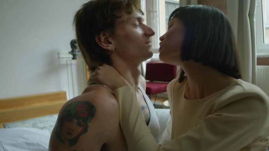 A man and a woman are sitting together on a bed and she has her hand around his neck and they kiss