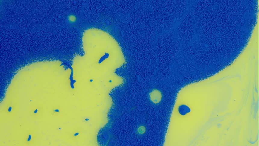 A blue substance moves and yellow emerges as blue pieces move over it as it spreads and colors mix around the edges