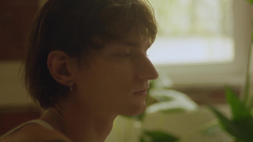 A young man sitting in a room with a gentle breeze blowing his hair and leaves on a house plant