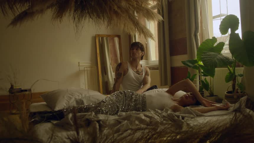 A man sits on a bed next to a woman lying beside him with her arm across her forehead