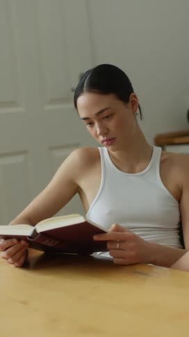 A young woman in a tank top reads a book while sitting at a desk indoors
