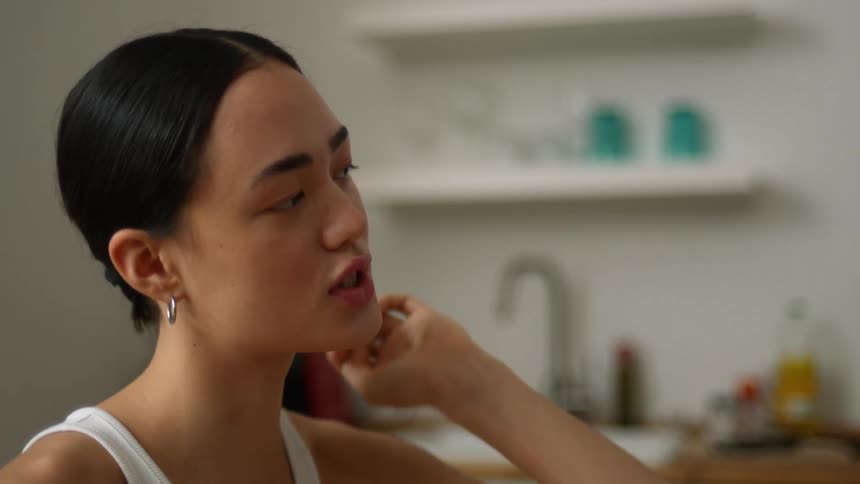A woman is sitting at a kitchen table with her hand on her chin and she speaks to someone