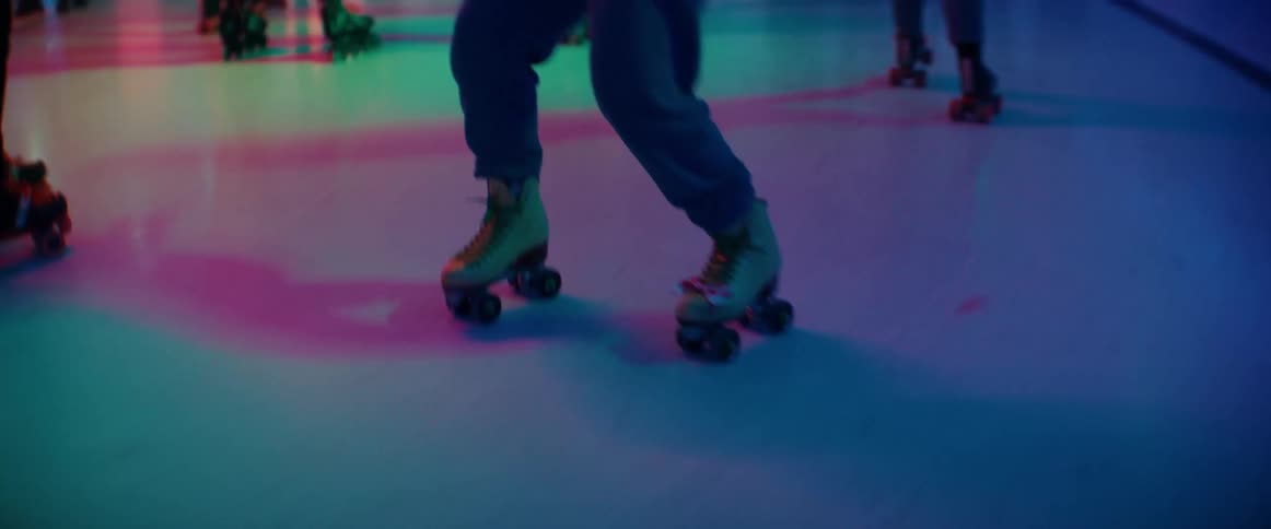Woman with wavy hair necklace white top and jeans dances while roller skating among crowd of roller skaters indoors
