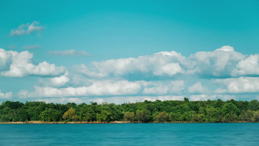 Cumulus clouds move across the sky over a forest of trees as water flows in the river and boats sail
