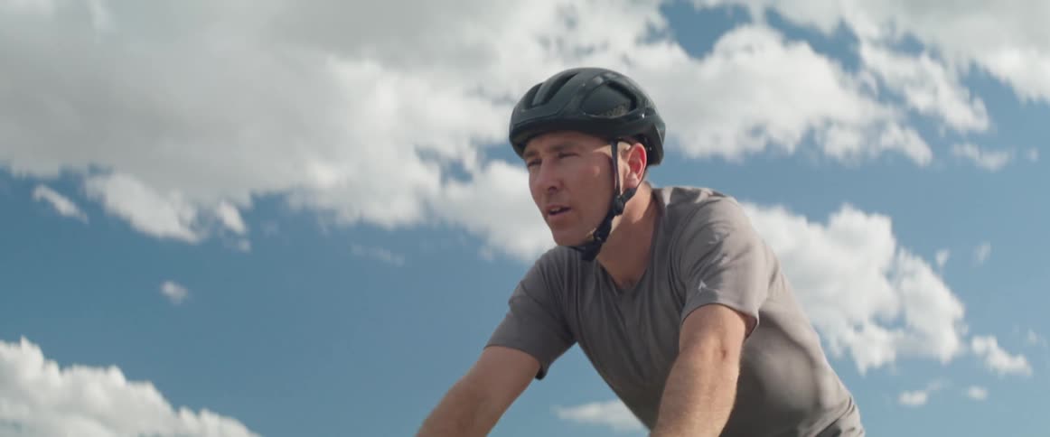 Mature man wearing a helmet rides a bicycle on a rural road at daytime under a blue sky