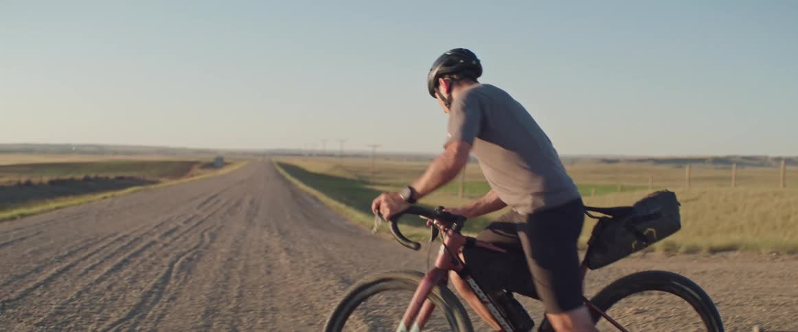 A man on a bike looks down a gravel road in a rural area before turning on the road and cycling down it