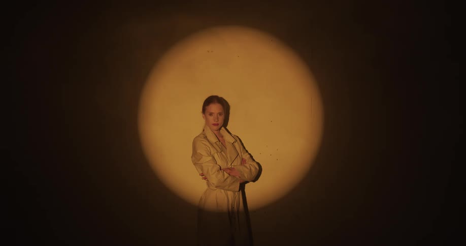 Woman with ponytail and brown coat stands with crossed arms in spotlight