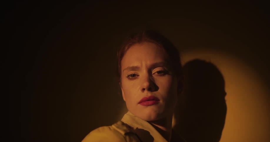 A woman wearing red lipstick is standing near a wall as a spotlight shines upon her she looks to her side and leans her head against the wall
