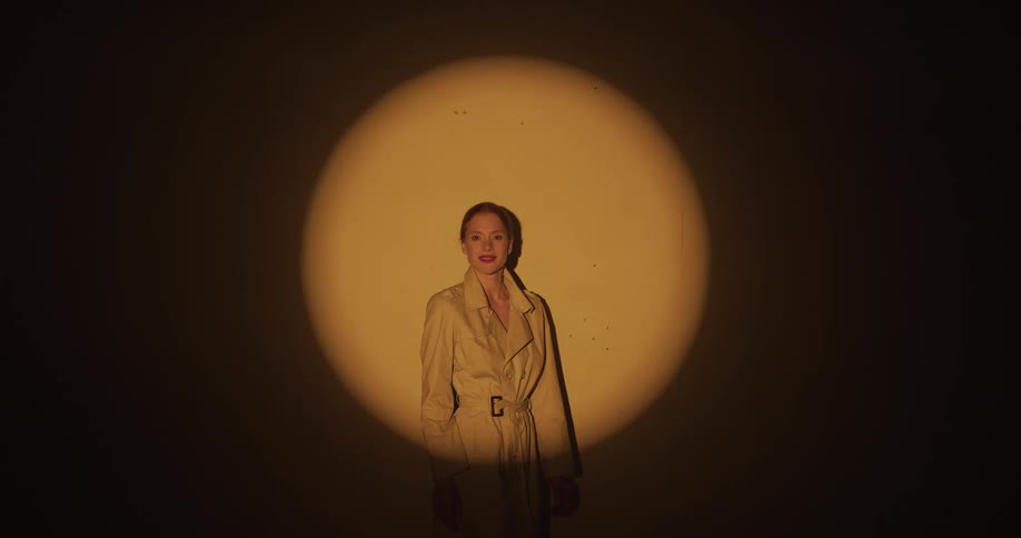 Female model with ponytail and brown coat stands in spotlight looking at camera in dark room