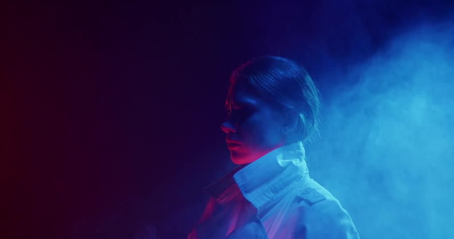 Light shines on a woman as smoke rises behind her as she stands in the darkness