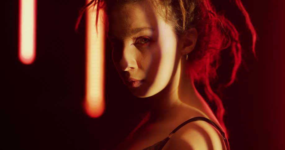 Young woman with a chaotic hairdo stands on a room surrounded by floating lights looking at the camera