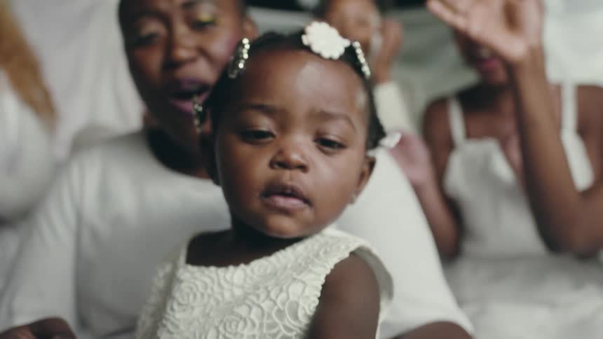 Close up of a toddler wearing white with woman cheering behind her