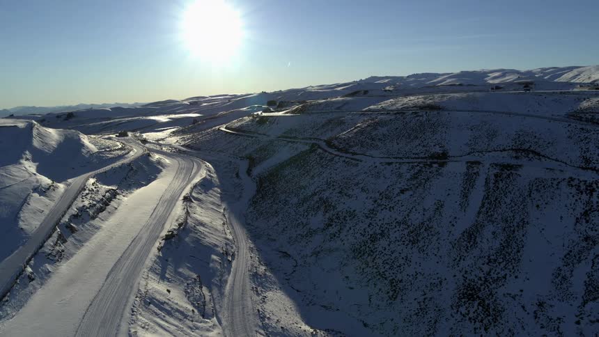 The sun shines brightly upon snow covered roads wrapped around mountains under a clear sky