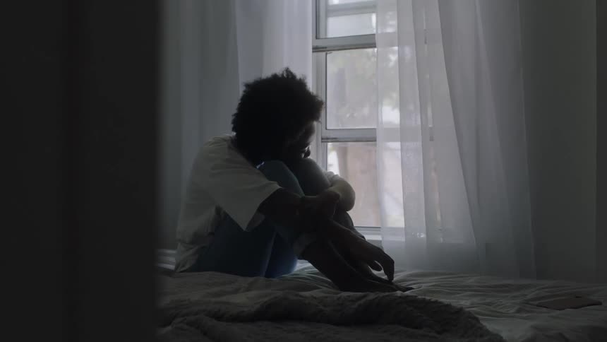Thoughtful and sad black woman sitting on the bed looking through the window
