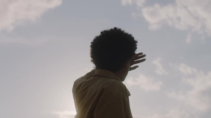 Young black man standing outdoors covering his face against the sun with his hands