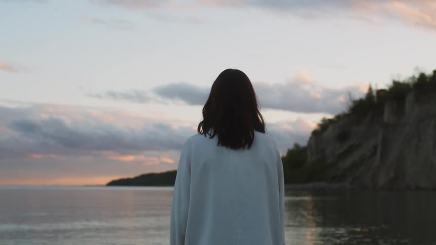 Woman stands on shore looking out towards horizon at dusk