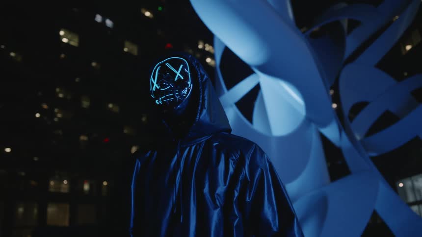 Person wearing hoodie and digital mask stands near large sculpture outside skyscraper at night