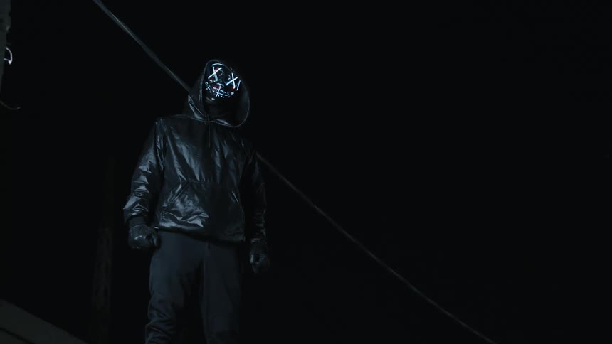 A person wearing all black and a mask stands in the dark