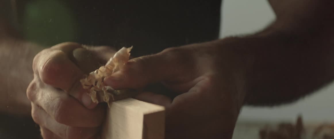 Hands move a small hand planer over the edge of a wooden plank