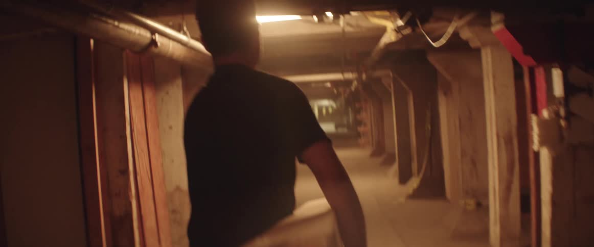 A man struggles a bit carrying a large heavy piece of lumber down a low ceilinged corridor