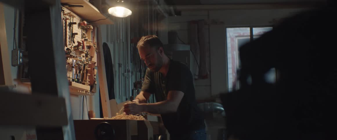 Carpenter uses a plane to file the side of a piece of wood over a workbench at night