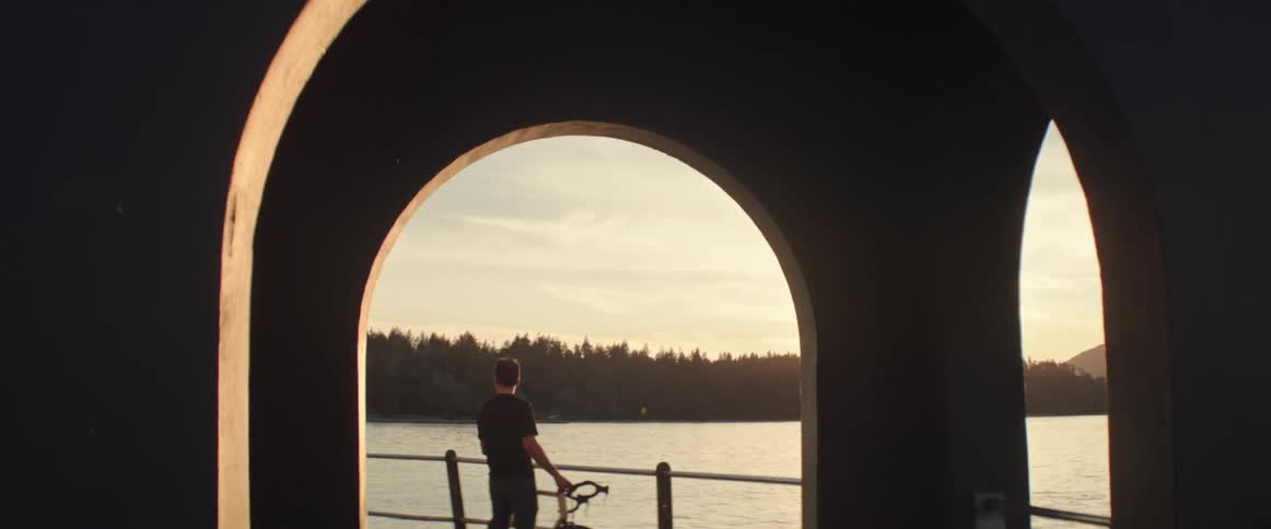 Man stands near rail with bicycle overlooking water and forest as sun shines from horizon