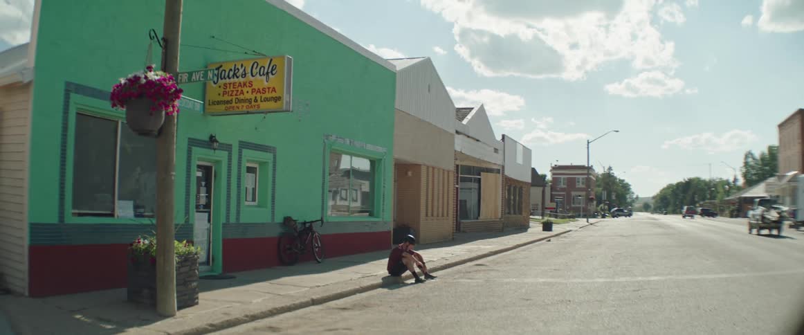 Male cyclist with helmet red shirt and black shorts sits on sidewalk drinking water from bottle and resting in town