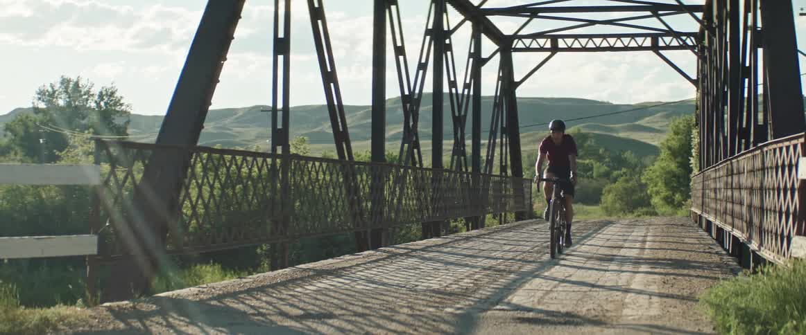 Male cyclist with helmet red shirt and black shorts rides bicycle across the bridge over the river