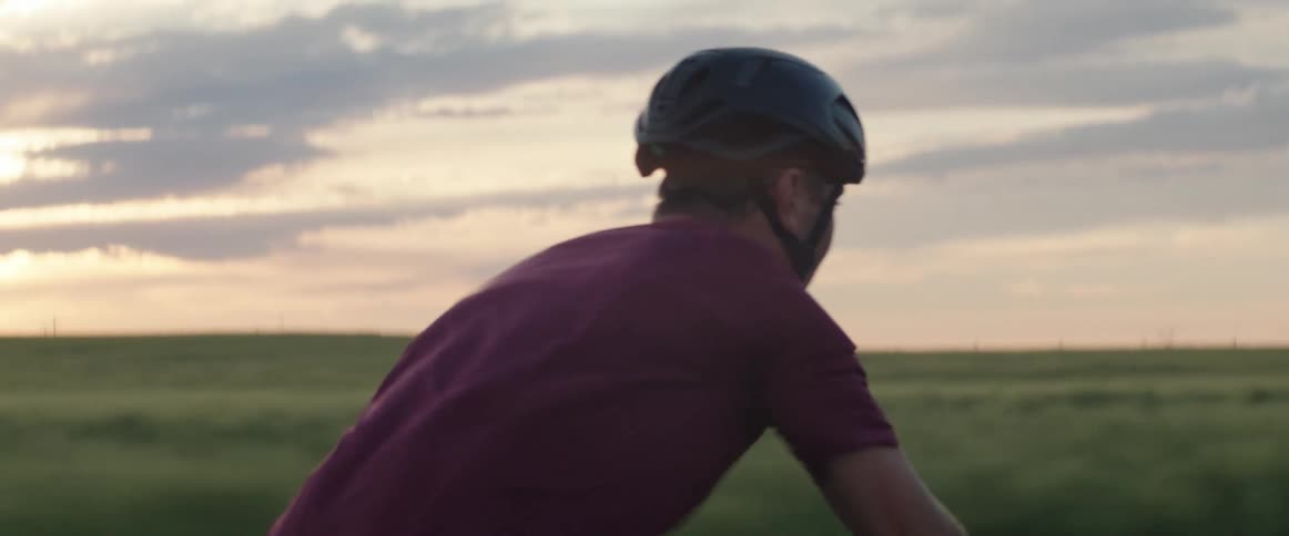 A man wearing a helmet is riding a bicycle on a rural road under a cloudy sunset sky