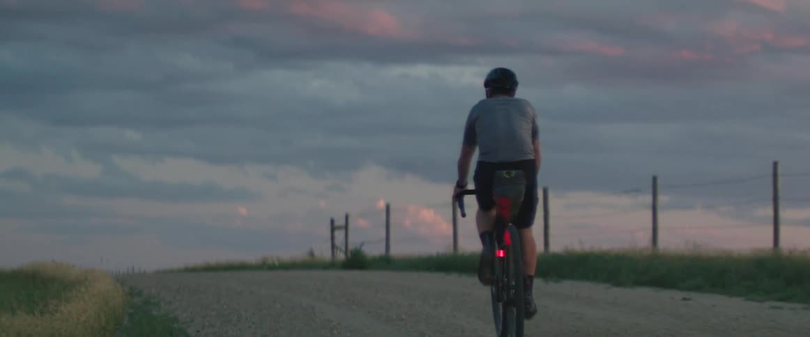 A man wearing a helmet is riding a bicycle on a rural road under a cloudy sky