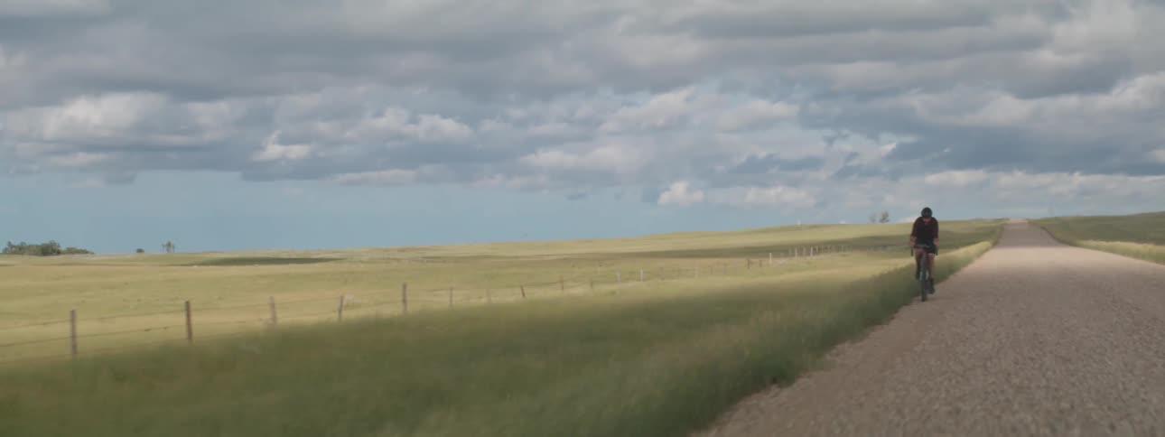 A man is riding a bicycle and pedaling hard on a wide open rural dirt road