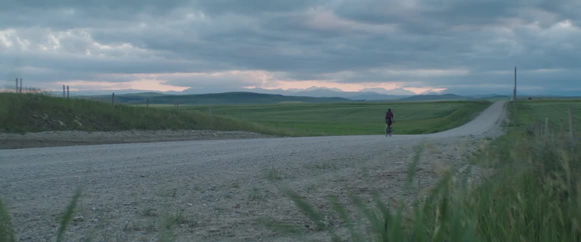 A person rides a bicycle on a rural dirt road towards distant foothills