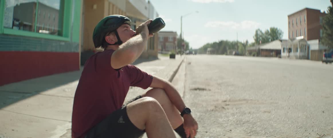 Man wearing a bike helmet drinking from his water bottle while sitting on the sidewalk in a small town