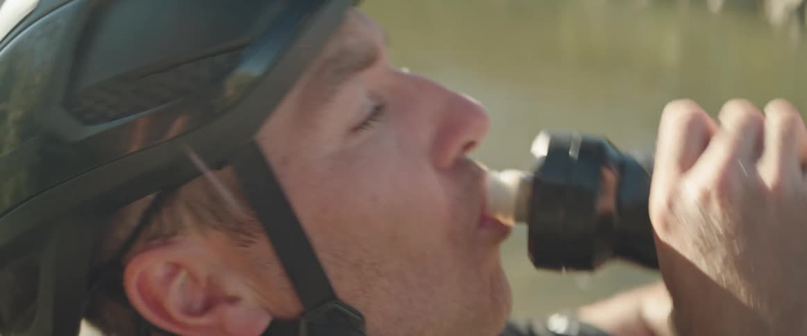 Male cyclist with black helmet and t shirt sits by stream drinking water from bottle and getting up