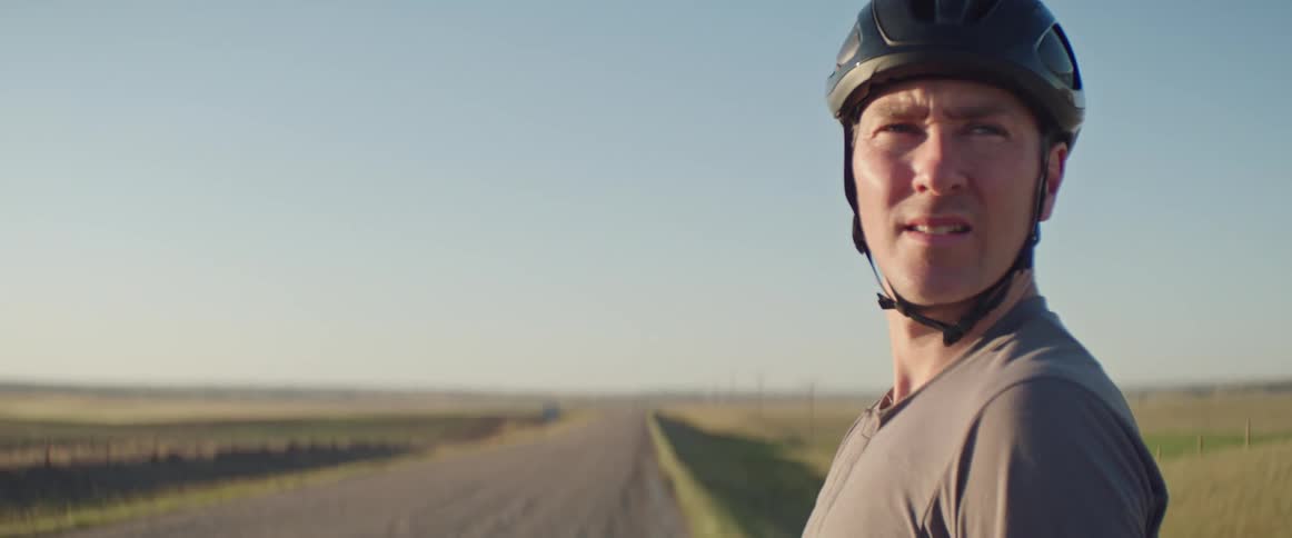 Man wearing a bicycle helmet waits along his bike on the side of a dirt road in a rural area