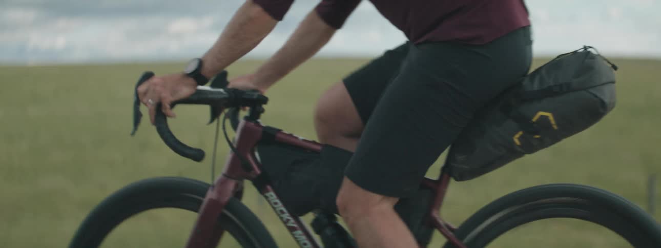 Man with red sweatshirt black shorts socks and shoes rides bicycle on countryside next to green field
