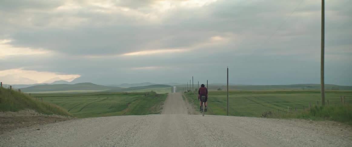 A person rides a bicycle on a rural dirt road towards the mountains